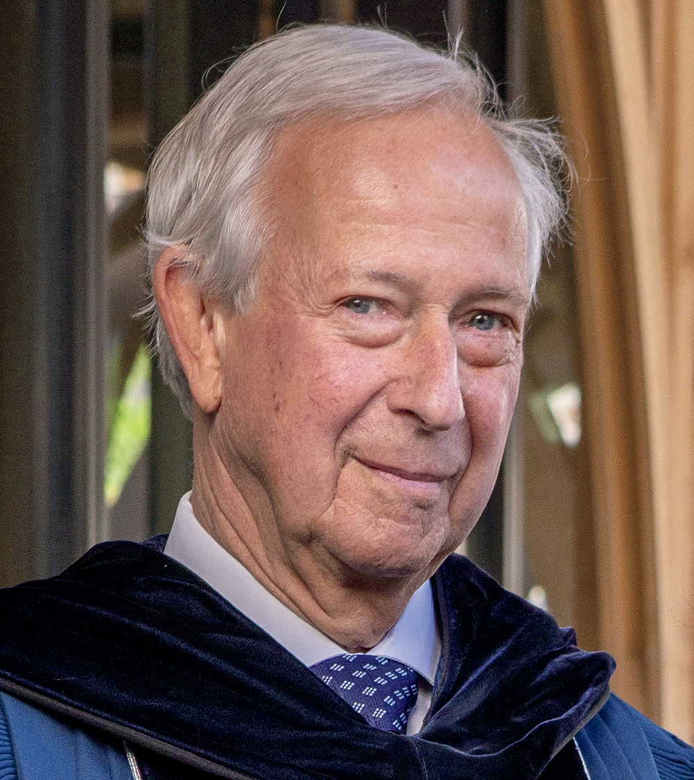 A headshot of Richard H. Brodhead wearing a commencement gown for graduating doctoral students. Brodhead has pale skin, blue eyes, and white hair, and he smiles at the viewer. 