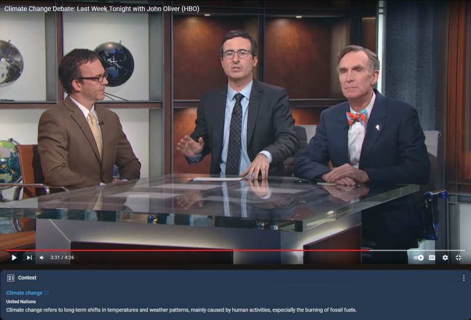 A screenshot from the show Last Week Tonight with John Oliver. John Oliver, a man with pale skin and short dark hair, wears a gray suit, blue collared shirt, and gray tie. He faces the audience and sits between Bill Nye and a person described as “a climate denier.” Bill Nye, a man with pale skin and short gray hair, wears a blue suit, a white collared shirt, and an orange and blue tie. He calmly faces the audience with his hands folded on the table. The climate denier, a man with pale skin and short dark ha