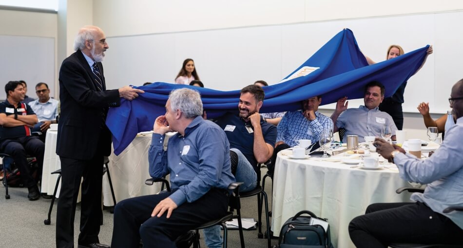 A photo of a role-playing event to educate the public on climate change issues. A man with white hair and a white beard wears a black suit and faces a group of people seated at tables. He holds one side of a large blue cloth that stretches over the heads of the people seated. A person stands behind the people at the tables and holds the other side of the cloth above their heads.