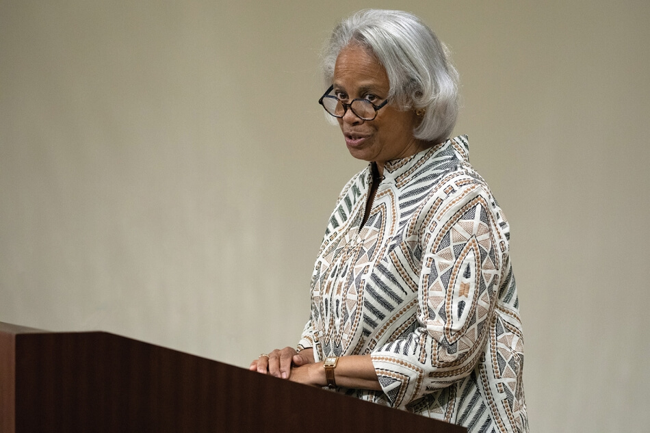 A photo of Ann Fudge, a woman with brown skin and white shoulder-length hair. She wears a white, black, and gold coat with the zipper on the collar. Photo by Martha Stewart Photography.