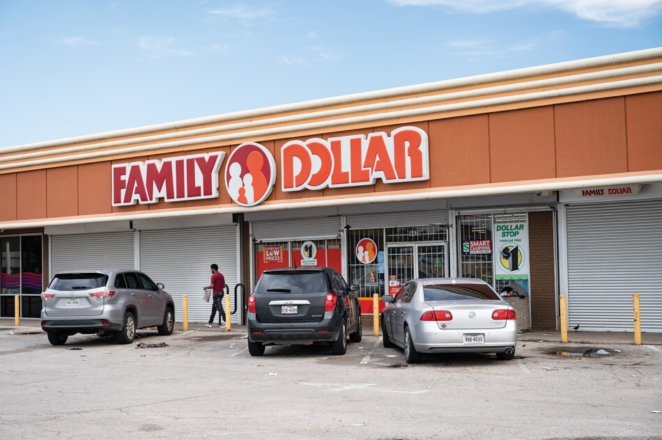 A photo of a pedestrian on the sidewalk between parked cars and a shuttered storefront for a Family Dollar in Texas. Photo by Cindy Elizabeth.