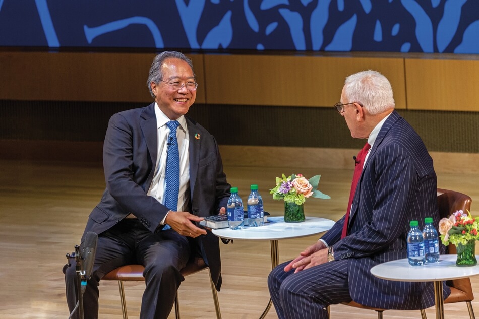 Yo-Yo Ma and David M. Rubenstein sit smiling and facing each other onstage during the 2022 Induction ceremony. Ma has brown skin and short gray hair, and wears wire glasses, a bright blue tie, a white collared shirt, a dark blue suit, and a lapel pin in support of the UN Sustainable Development Goals. Rubenstein has pale skin and short white hair. He wears plastic glasses, a red tie, a white collared shirt, and a blue pinstriped suit. Photo by Michael DeStefano Photography.
