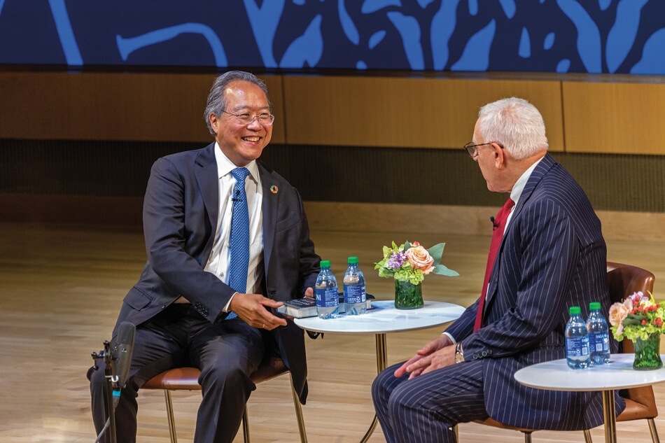Photo by Michael DeStefano Photography. Yo-Yo Ma and David M. Rubenstein sit smiling and facing each other onstage during the 2022 Induction ceremony. Ma has brown skin and short gray hair, and wears wire glasses, a bright blue tie, a white collared shirt, a dark blue suit, and a lapel pin in support of the UN Sustainable Development Goals. Rubenstein has pale skin and short white hair. He wears plastic glasses, a red tie, a white collared shirt, and a blue pinstriped suit. Photo by Michael DeStefano Photography.