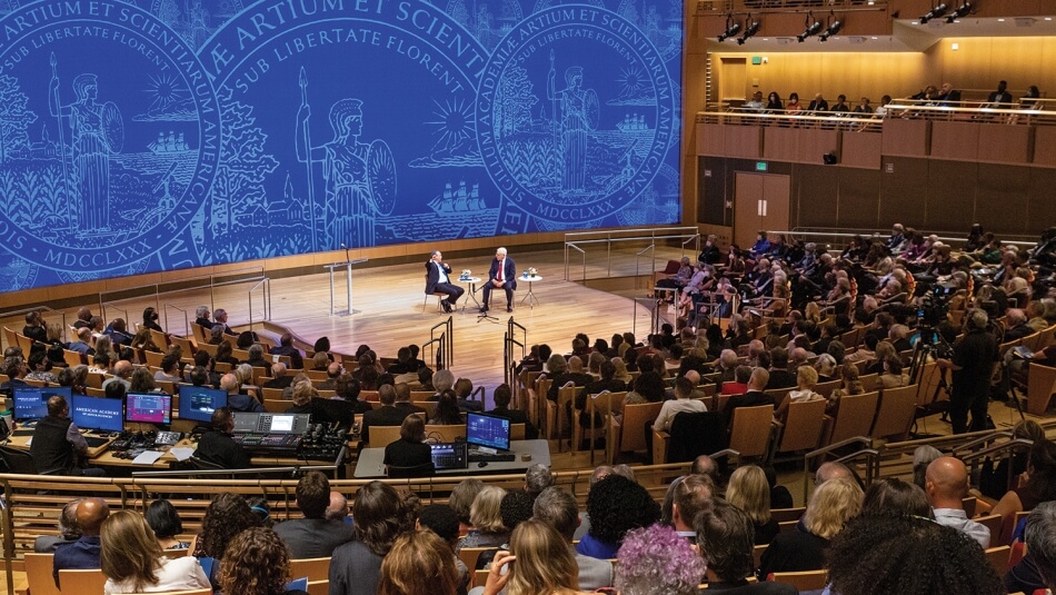 A wide shot of Yo-Yo Ma and David M. Rubenstein, taken from the audience during the 2022 Induction ceremony. Ma and Rubenstein sit facing each other onstage. Ma gestures one hand toward the audience while Rubenstein looks on. Photo by Michael DeStefano Photography.