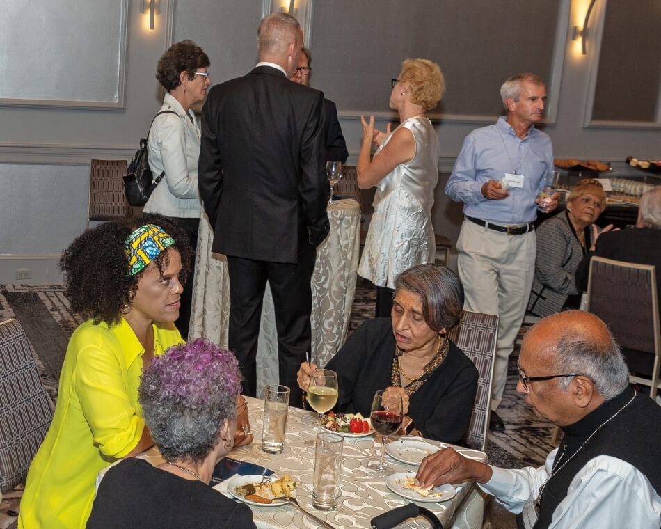 People at a reception. Some standing and some seated, eating, drinking, and talking. 