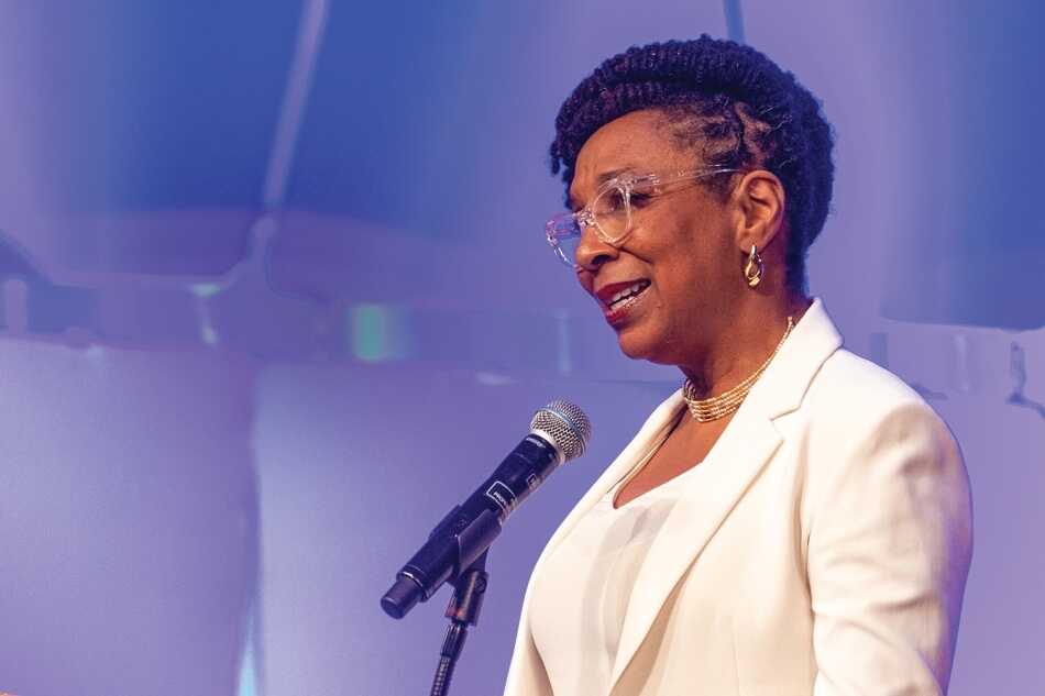 Photo by Michael DeStefano Photography. A photo of Kimberlé W. Crenshaw standing at a microphone at the 2022 Induction ceremony. She has brown skin and long black hair, worn in braids done up in an elegant twist. She wears a white suit, gold earrings, and a gold necklace. She smiles and addresses the audience. Photo by Michael DeStefano Photography.