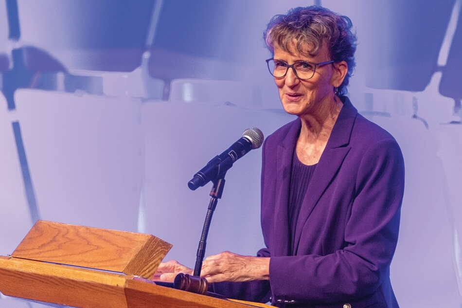 A photo of Mary Kay Henry standing at a podium at the 2022 Induction ceremony. She has pale skin and short gray hair. She wears glasses, a purple blouse, a purple suit, and metallic earrings. She smiles and addresses the audience. Photo by Michael DeStefano Photography.