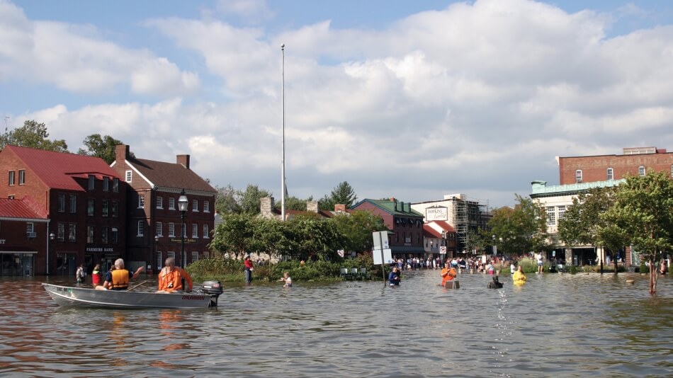 A boat floats down a body of water that has filled a space that was once a street. Storefronts, houses, and trees stand in the background, flooded, and people wade through the water to reach each other.