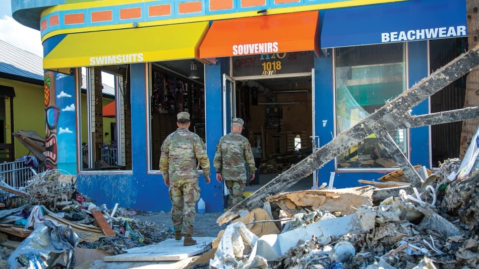Two people wearing army camouflage approach a decimated storefront in a building that is painted bright colors but is otherwise destroyed. Debris is scattered on either side of the path they walk to reach the door. 