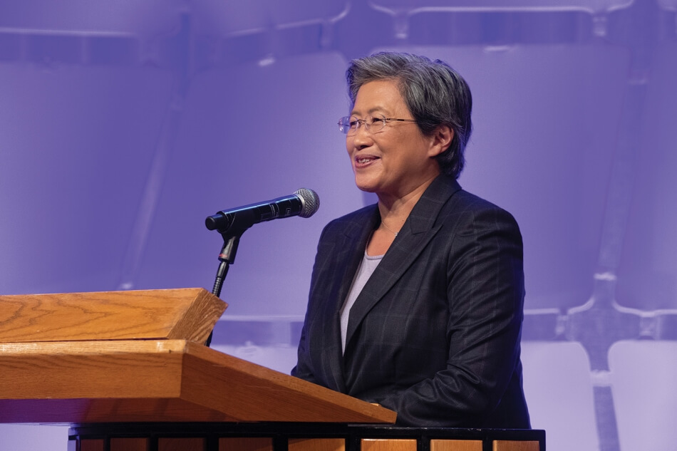 Photo by Martha Stewart Photography. A photo of Lisa T. Su standing at a podium at the 2022 Induction ceremony. She has brown skin and short gray hair, and wears wire glasses, a pale blouse, and a dark suit. She addresses the audience and smiles. Photo by Martha Stewart Photography.