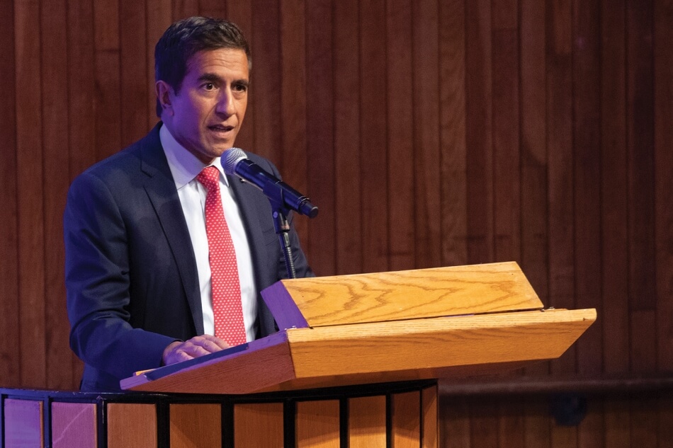 A photo of Sanjay Gupta standing at a podium at the 2022 Induction ceremony. He has brown skin and short black hair, and wears a white collared shirt, a coral tie, and a dark suit. He addresses the audience. Photo by Martha Stewart Photography.