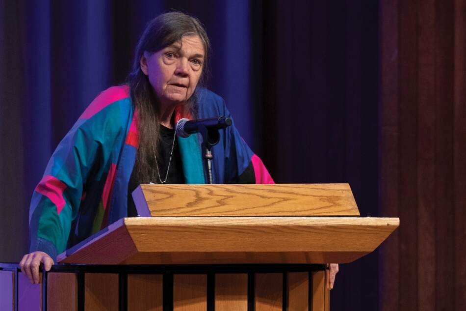 Photo by Martha Stewart Photography. A photo of Patricia Limerick standing at a podium at the 2022 Induction ceremony. She has pale skin and long dark hair. She wears a multicolored suit, black blouse, and a silver necklace. She addresses the audience. Photo by Martha Stewart Photography.