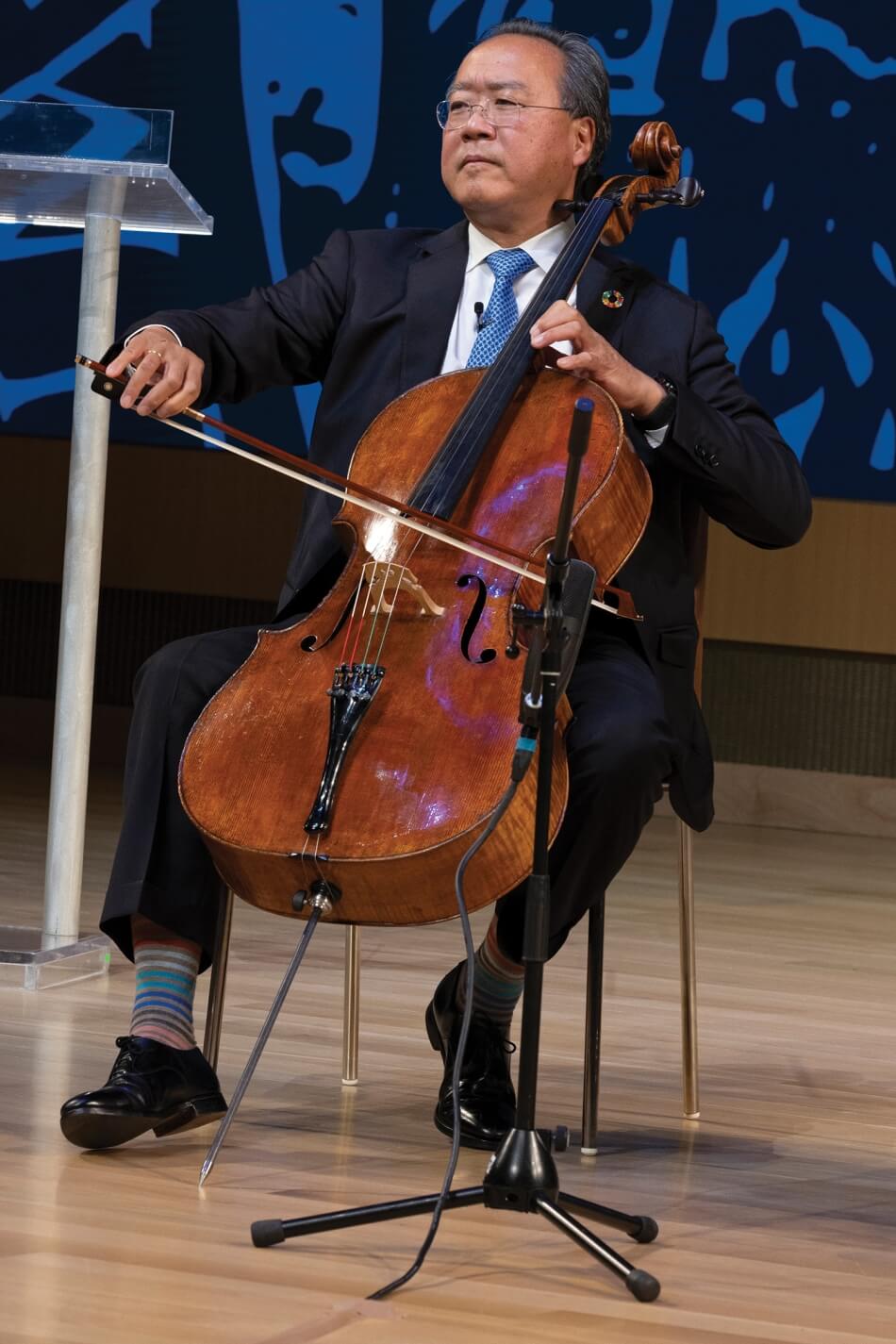 A portrait of Yo-Yo Ma playing the cello during the 2022 Induction ceremony. He has brown skin and short gray hair, and wears wire glasses, a bright blue tie, a white collared shirt, a dark blue suit, multicolored striped socks, black shoes, and a lapel pin in support of the UN Sustainable Development Goals. He holds a bow to the strings of a cello in one hand and balances the neck of the cello with the other. Photo by Martha Stewart Photography.