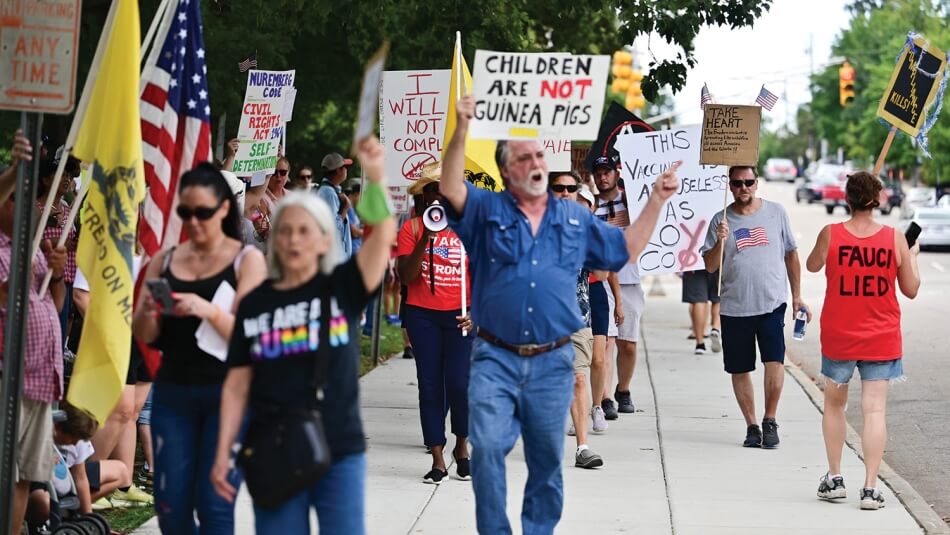 An interior image from the fall 2023 issue of Dædalus. Protestors at a “freedom of choice” rally in September 2021 pace on a sidewalk and some walk in the streets holding handwritten signs or wearing shirts with text on them, some of which read: “Children are not guinea pigs,” “Fauci lied,” and “I will not comply.” Photo by Anthony Crider. Published under a Creative Commons Attribution-NonCommercial 2.0 Generic (CC BY-NC 2.0) license.