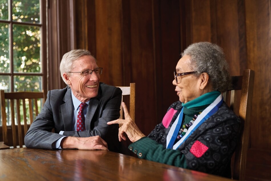 Photo by Noah Willman. A photo of David Oxtoby and Marian Wright Edelman sitting side by side at a wooden table. Both smile, and Edelman holds one hand up as if in midspeech. Photo by Noah Willman.