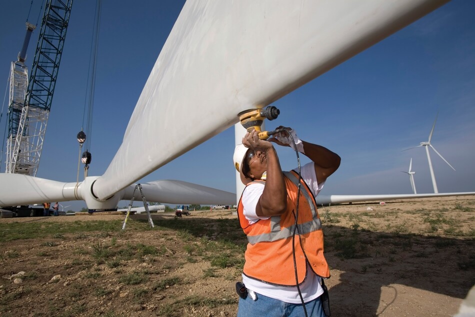 A construction worker holds a tool to the propeller of a wind turbine. The worker has black hair and brown skin, and wears an orange vest, safety goggles, and a white hard hat. The turbine stretches over a field of grass and beneath a crane. Two other wind turbines stand in the background.