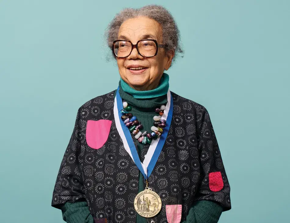 Photo by Noah Willman. A headshot of Marian Wright Edelman, an elderly woman with brown skin and curly gray hair parted in the middle, as she faces the camera and smiles. Edelman wears two blue turtlenecks, a black and gray sweater, and a colorful beaded necklace. Photo by Noah Willman.