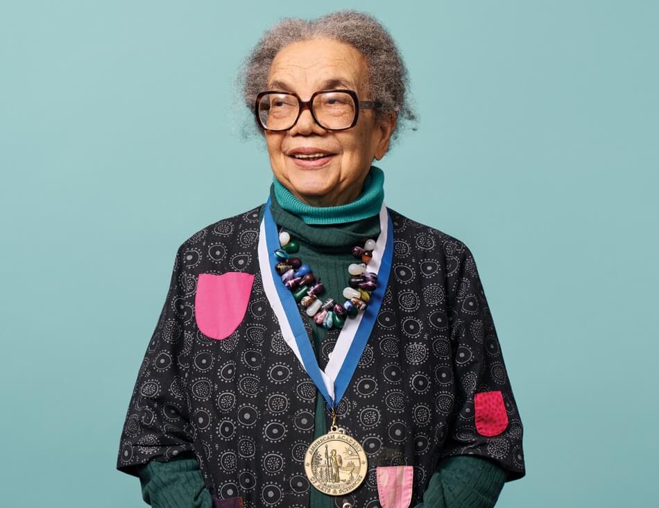 A headshot of Marian Wright Edelman, an elderly woman with brown skin and curly gray hair parted in the middle, as she faces the camera and smiles. Edelman wears two blue turtlenecks, a black and gray sweater, and a colorful beaded necklace. Photo by Noah Willman.