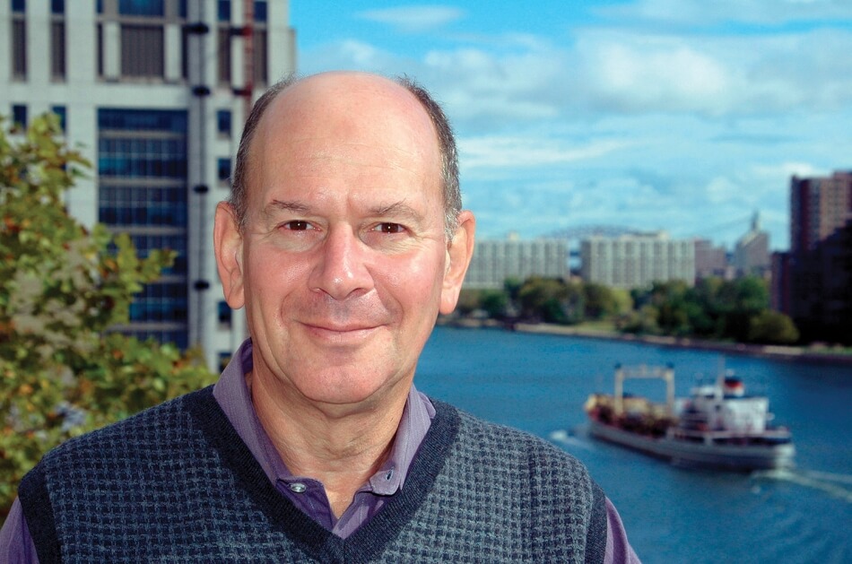 A portrait of Jesse H. Ausubel, a man with pale skin and graying hair. He faces the camera and smiles while standing in front of a river with buildings on both sides of its bank. Ausubel wears a blue and gray vest over a purple collared shirt. Photo courtesy of Jesse H. Ausubel.