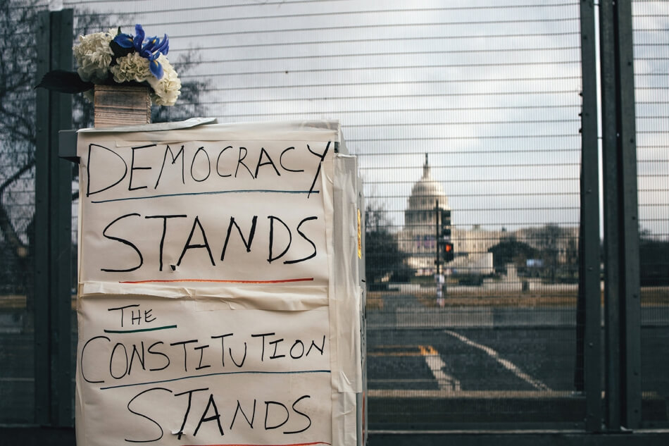 A close-up shot of a fence across the street from the Capitol Building in Washington, D.C. Flowers are pinned to the fence above a handwritten sign that reads: “Democracy stands, The constitution stands.” Photo by Brendan Beale on Unsplash.