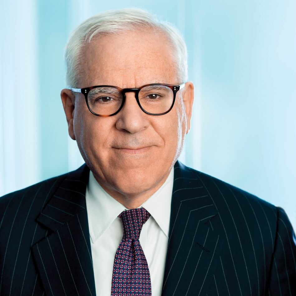 A headshot of David M. Rubenstein. He faces the camera and smiles. He has pale skin and short white hair. He wears plastic glasses, a blue and red tie, a white collared shirt, and a blue pinstriped suit. Photo courtesy of David M. Rubenstein.