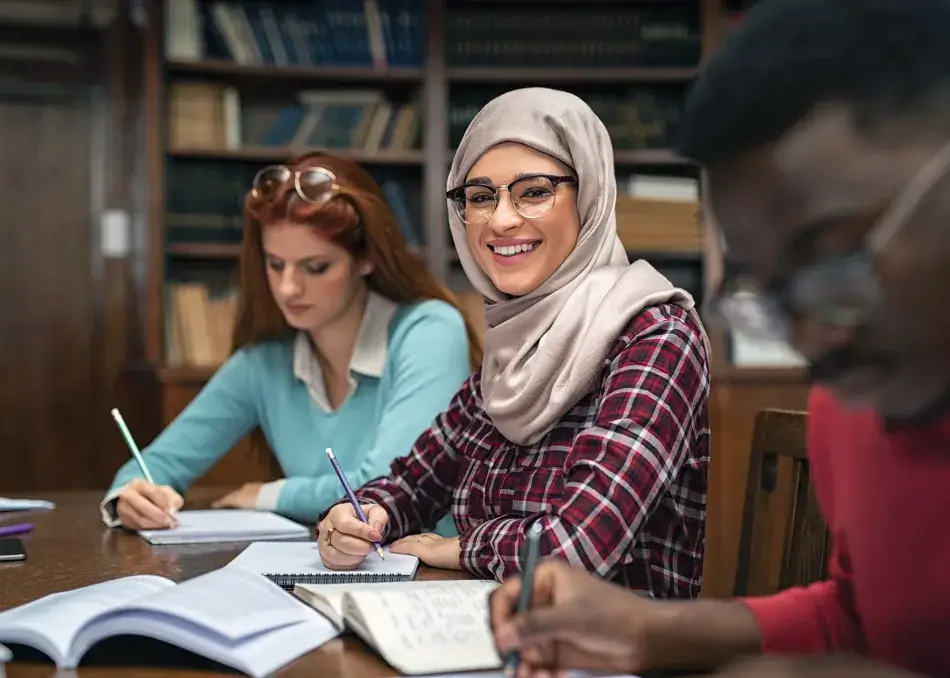 iStock.com/Ridofranz Happy young woman in hijab at university library looking at camera. Portrait of smiling female student wearing abaya and spectacles feeling confident. Islamic girl studying with multiethnic students.