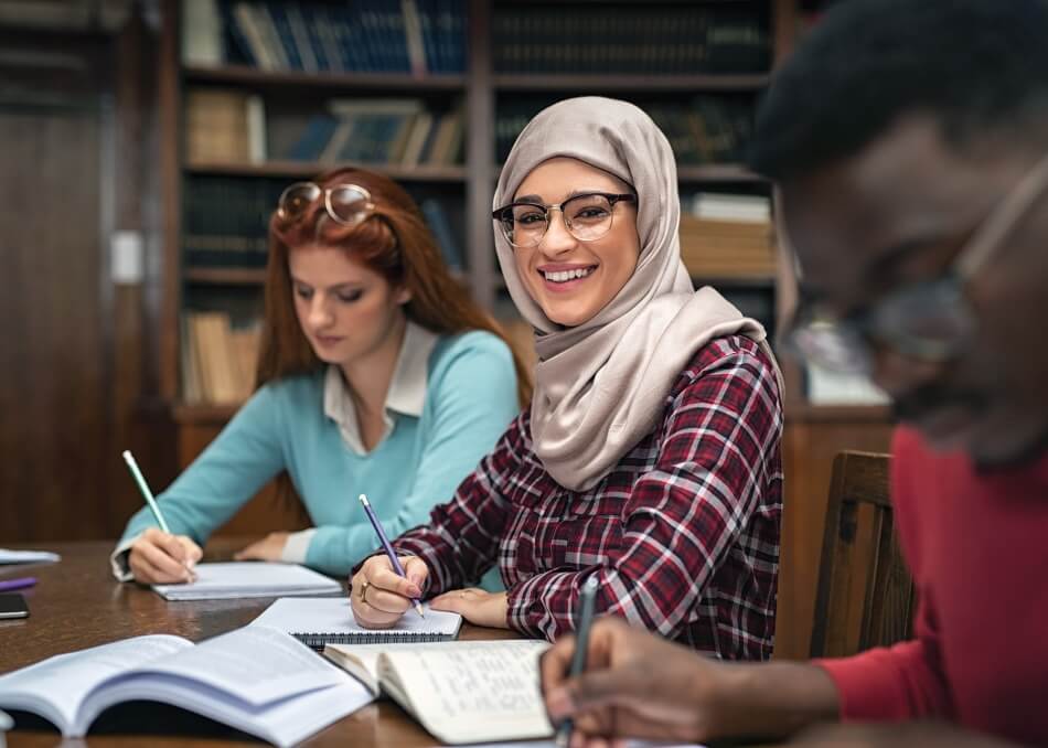 Happy young woman in hijab at university library looking at camera. Portrait of smiling female student wearing abaya and spectacles feeling confident. Islamic girl studying with multiethnic students.