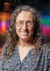 A headshot of Frances Hellman, a person with pale skin, blue eyes, and long curly graying hair. She wears business attire, faces the viewer, and smiles.