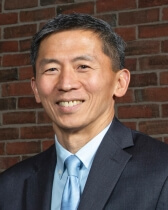 Headshot of Goodwin Liu, who has tan skin and short black hair. He wears a gray suit, blue tie, and faces the viewer smiling.