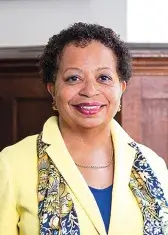 A headshot of Joanne Berger-Sweeney, a person with brown skin, brown eyes, and short curly black hair. She wears business attire, faces the viewer, and smiles.