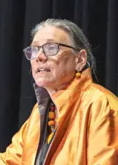 A photo of Laurie L. Patton, a person with light skin and light brown hair. She is wearing glasses and a orange blazer and is facing an unseen audience.
