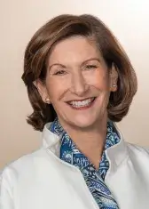 A headshot of Nancy Peretsman, a person with pale skin, brown eyes, and shoulder-length bronze hair. She wears business attire, faces the viewer, and smiles.