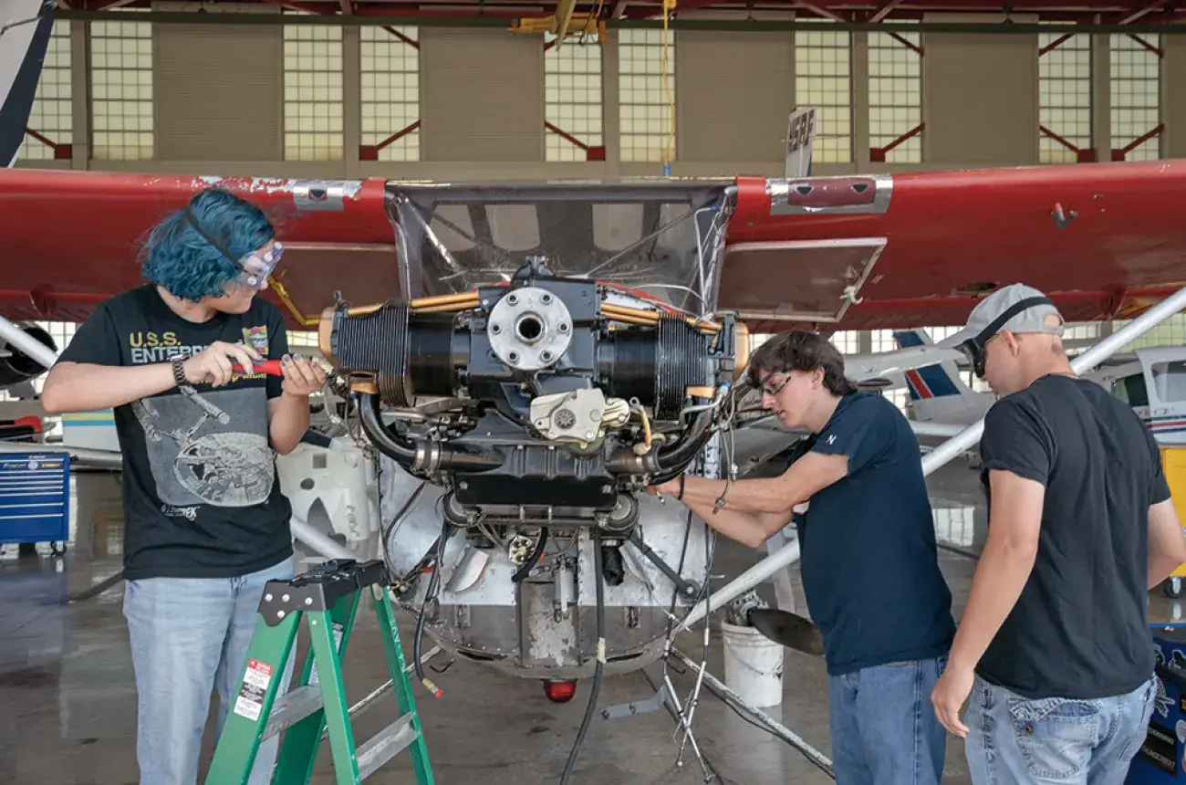Three people who are wearing protective goggles work on the front of an airplane that is red and white. Two are holding tools while one is looking at the plane. 