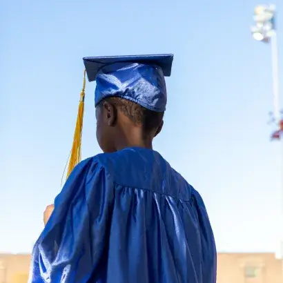 Young Black boy wearing a blue graduation cap and gown faces away from the camera outside.