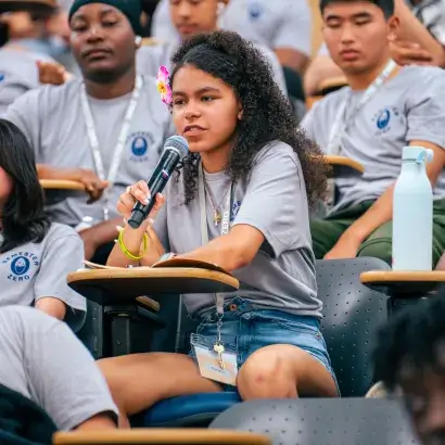 Students seated in a large room with ascending rows of desks, all wearing matching grey t-shirts. One girl is seated at a desk holding a microphone.