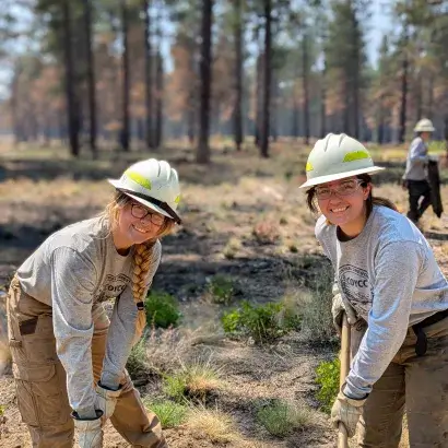 Two young women with hard hats and protective glasses in the woods holding long tools
