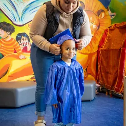 A mother attaching a graduation cap to a young child who is also wearing a graduation gown