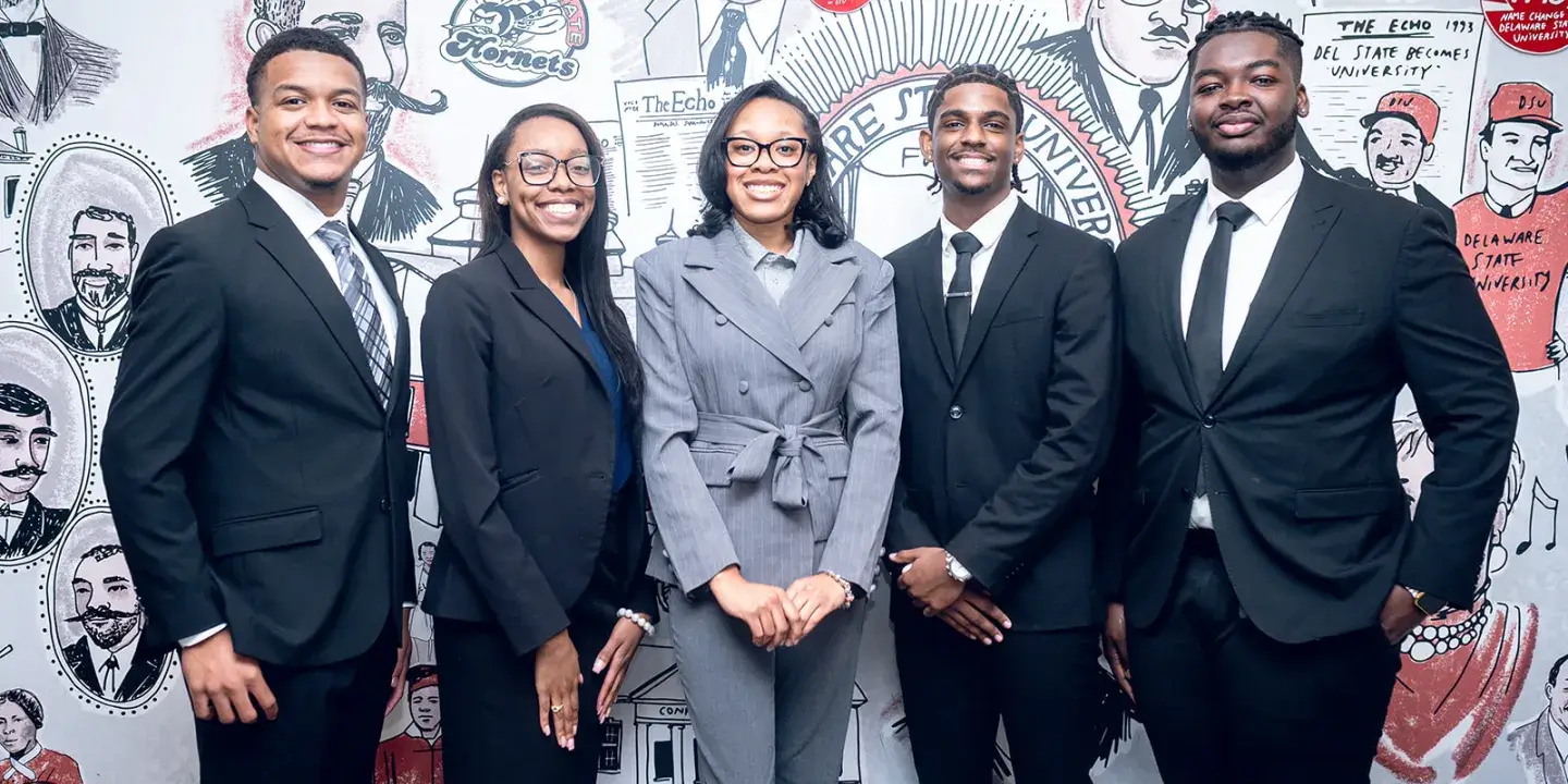 Group of five Black young adults in suits stand in front of a mural.