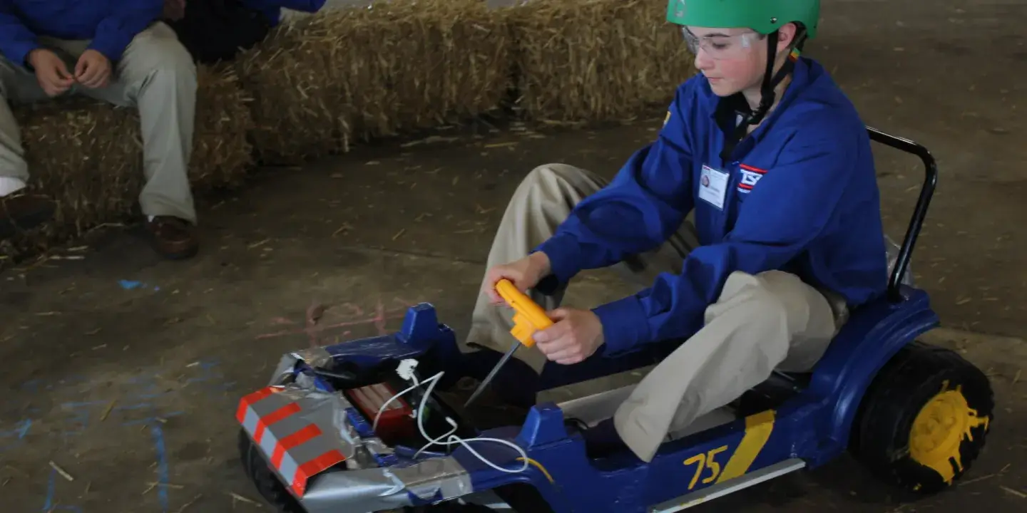 A student riding a homemade go-kart vehicle around a barn