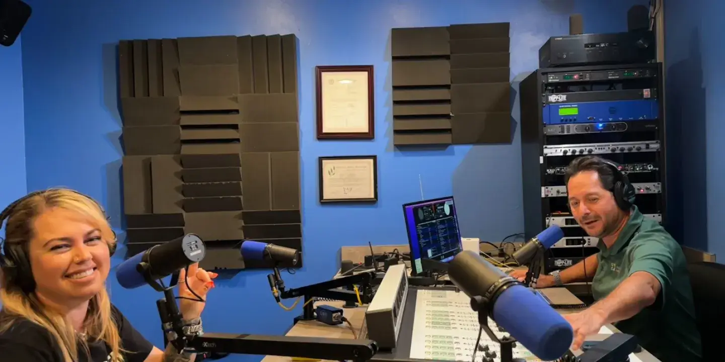Two people are seated in a room set up for a radio interview, posing and smiling for the camera 