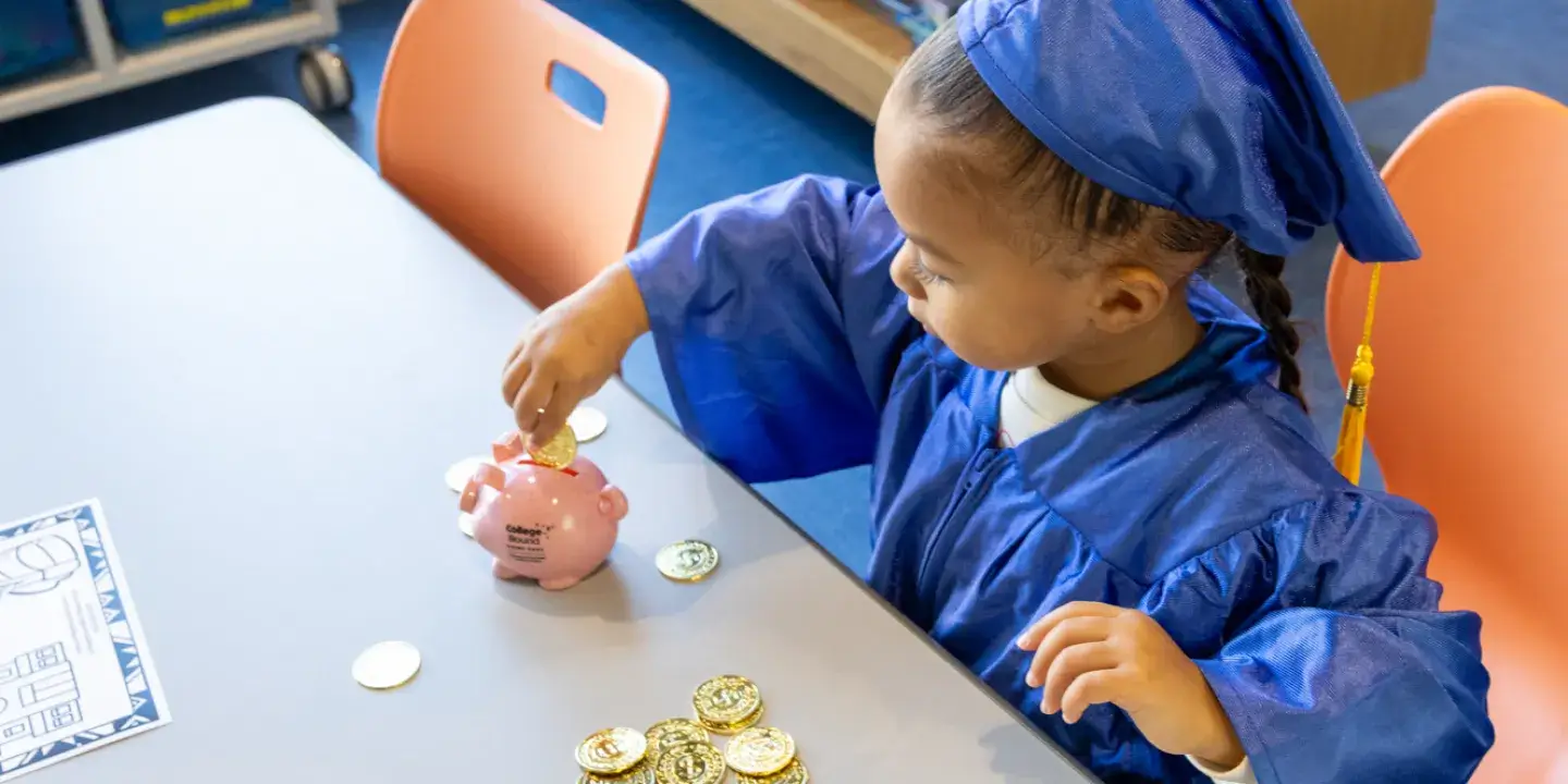 A young Black boy sitting at a table counting out coins with a blue graduation gown and cap on