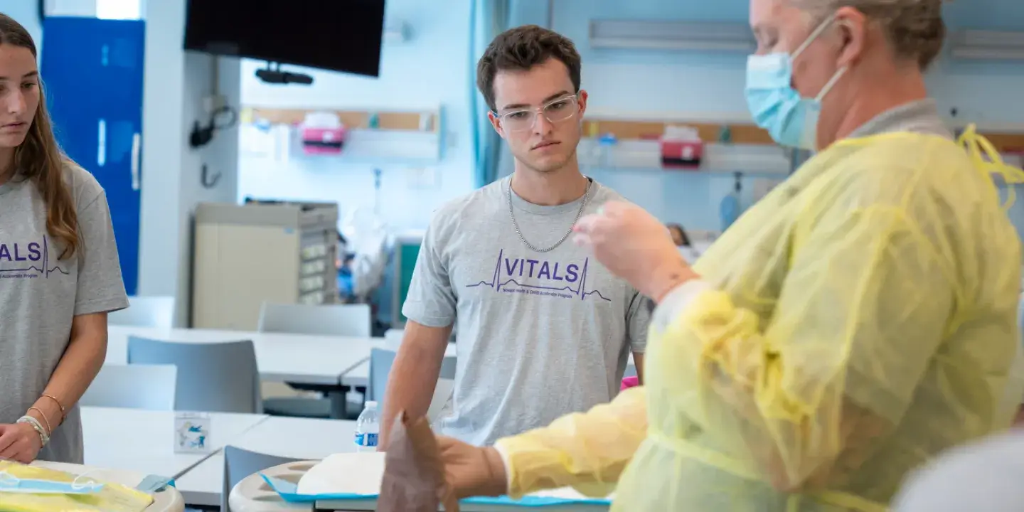 A nurse in a yellow cover teaching two students in a grey t-shirt 