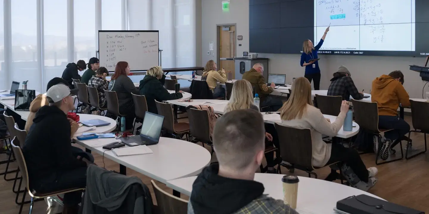 College math class with students seated at long tables facing a teacher pointing to an electronic whiteboard.