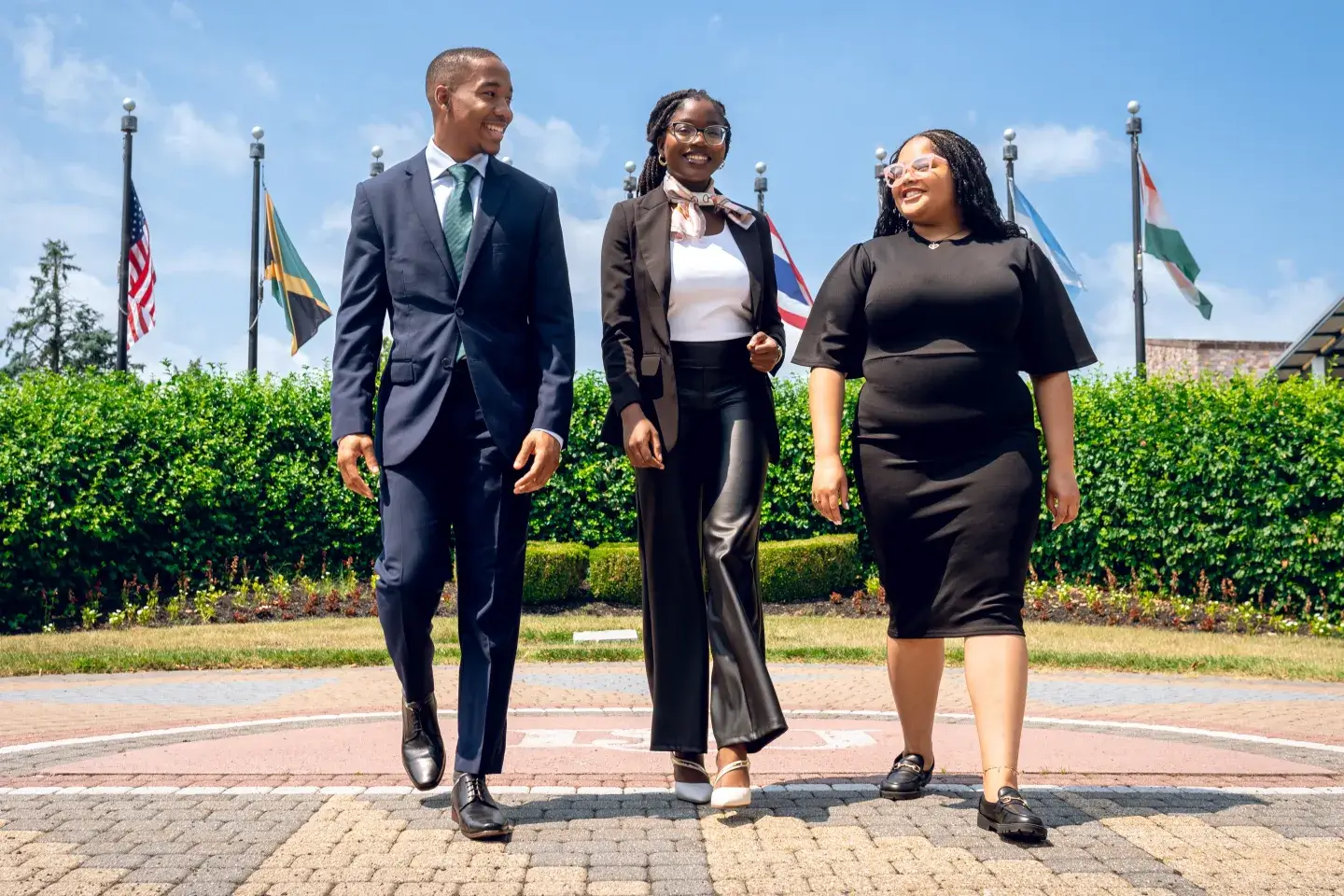 Group of three Black young adults in suits walking on an outdoor plaza in front of flags.