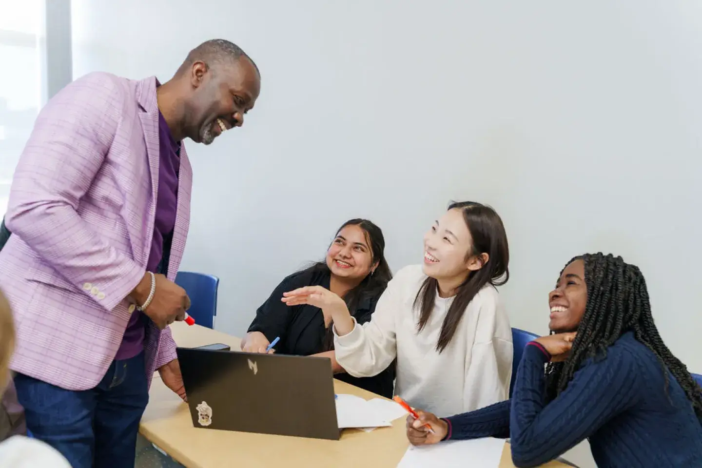 A Black male professor leans over to talk with three female students of different races who are smiling at him.