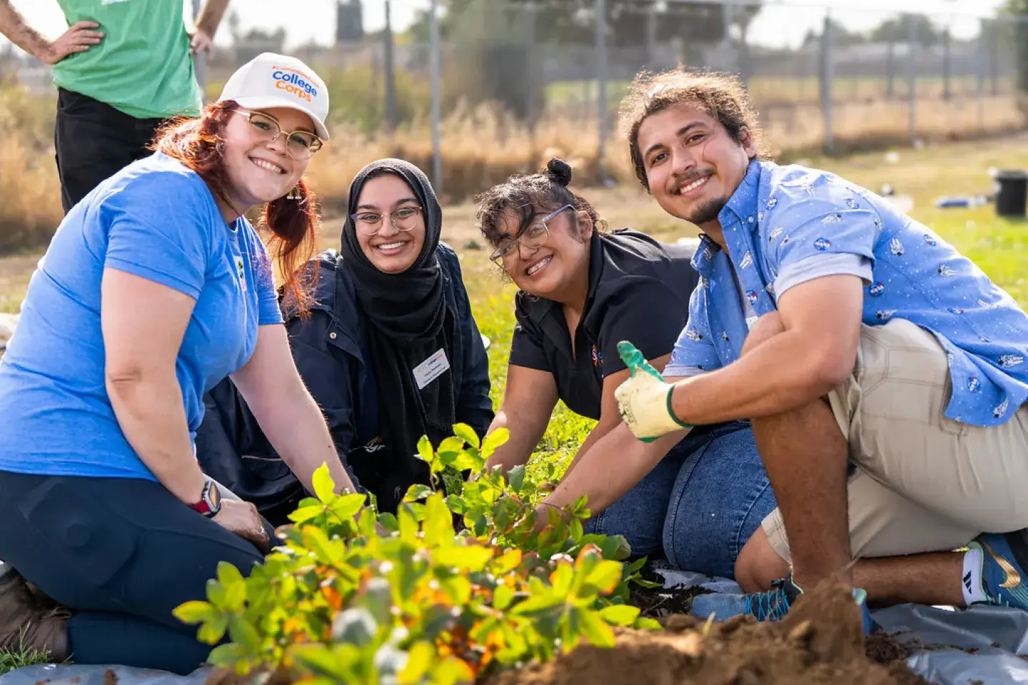 Four diverse college-aged volunteers kneel on the ground planting a garden.