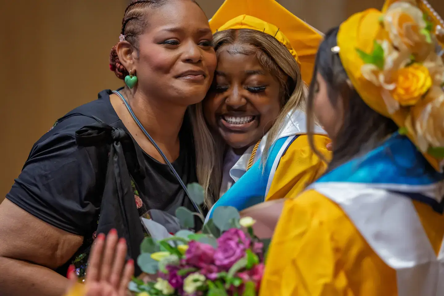 An older woman hugs a younger woman who has just graduated from high school. She is wearing a yellow graduation cap and gown. Both are teary eyed and smiling.