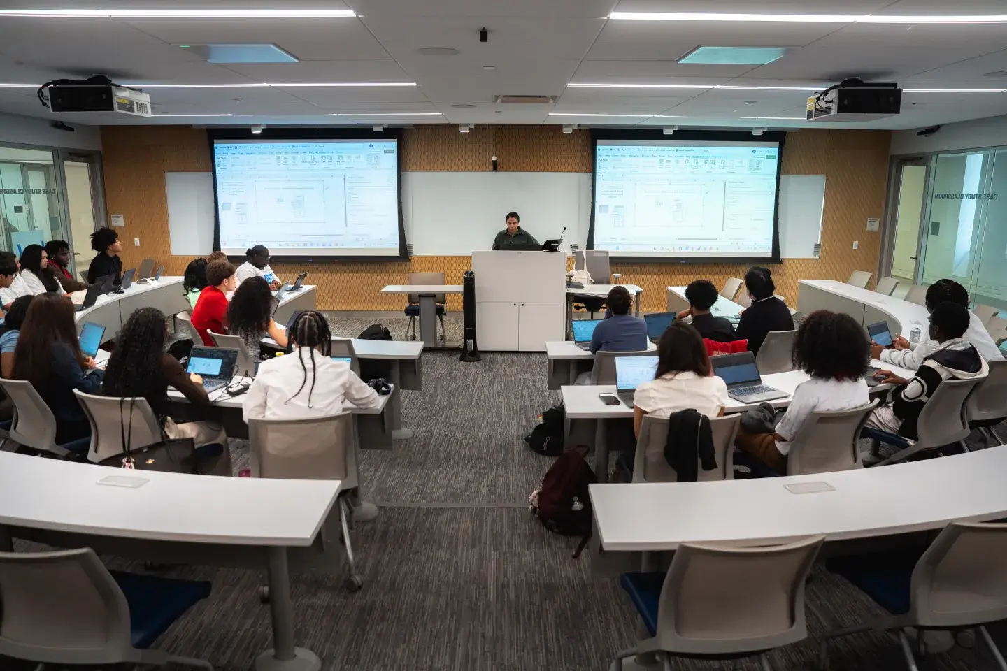 An auditorium style classroom featuring long, curved tables where students are sat. A teacher helms the front with two powerpoint presentations.