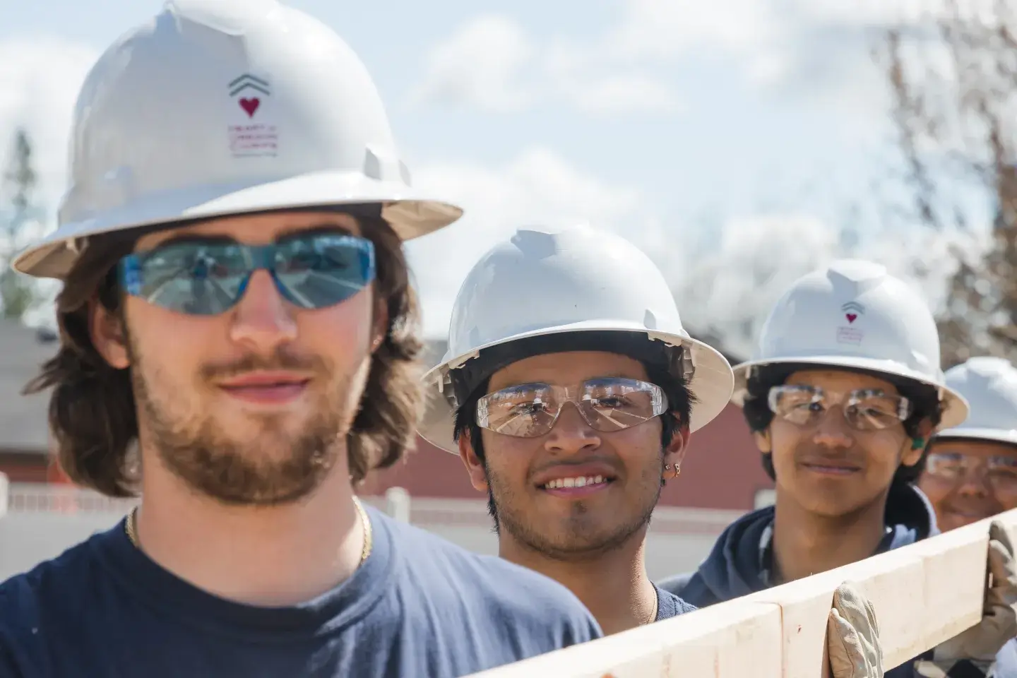 A group of young men in hard hats and protective eye glasses holding a large plank of wood