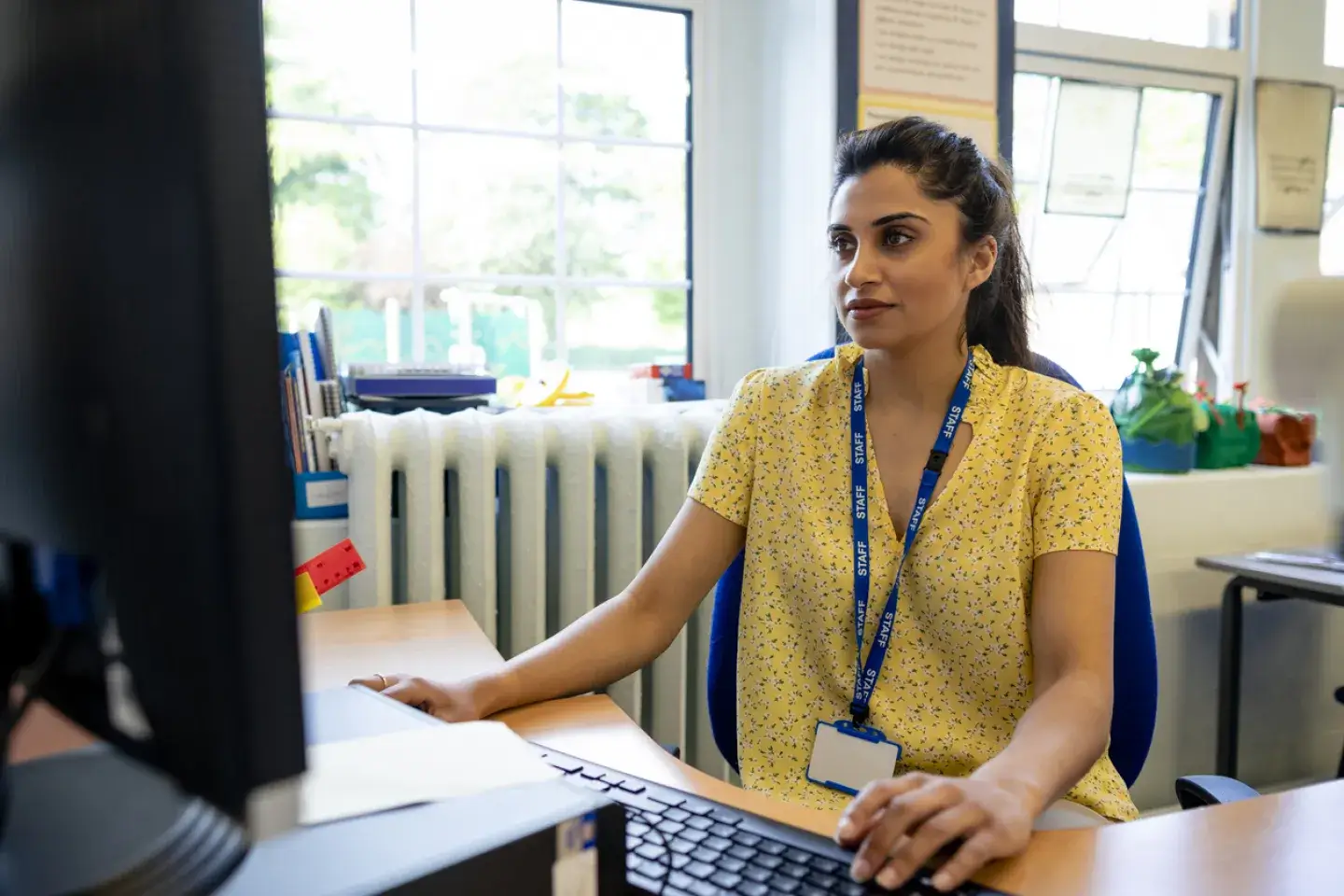 Teacher wearing a yellow shirt and lanyard using the computer at her desk